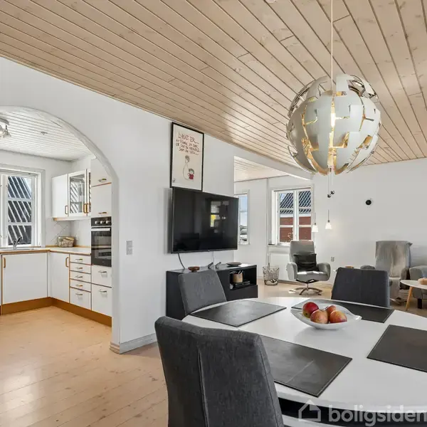 Bord står centralt dækket med glas og skålen med frugt i en stue med trægulve og loft, åbent til køkkenet gennem en bue. Gråt møblement omkranser rummet.