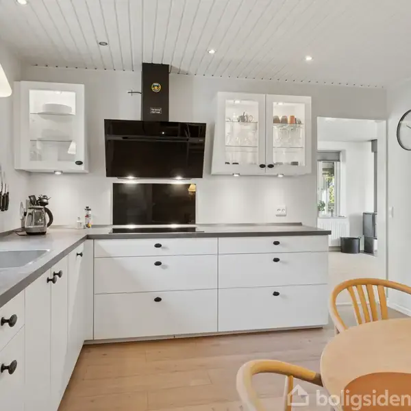 Et lyst køkken med hvide skabe og skuffer. Kanden står på bordpladen ved siden af en knivblok. Der er et bord med appelsiner. En vægur viser klokken halv tolv.