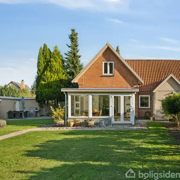 Et hus med rødt tag står i en have med velplejet græsplæne. Foran huset er en glaseret veranda. Til venstre ses en flagstang og nogle træer.