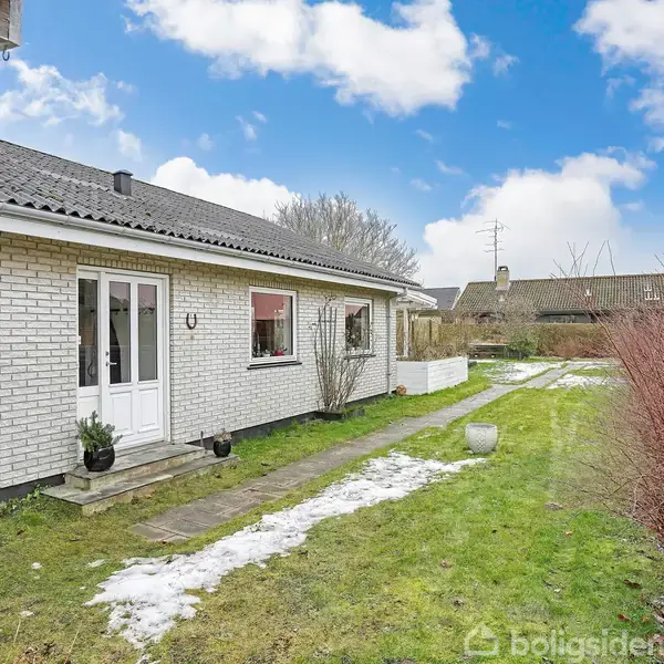 Hvid murstenshus med sorte tagsten, står ved siden af en græsplæne med sporadisk sne. Stensti fører til dobbeltglasdør, omgivet af buske og planter under en blå himmel.