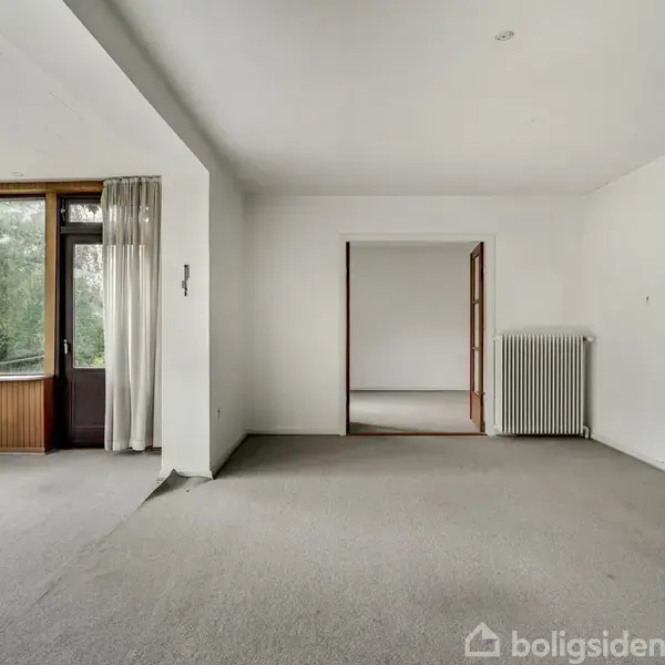 Rummet har hvide vægge og gråt gulvtæppe, store vinduer med gardiner mod venstre, og en radiator på højre væg. En døråbning fører til et tilstødende rum.