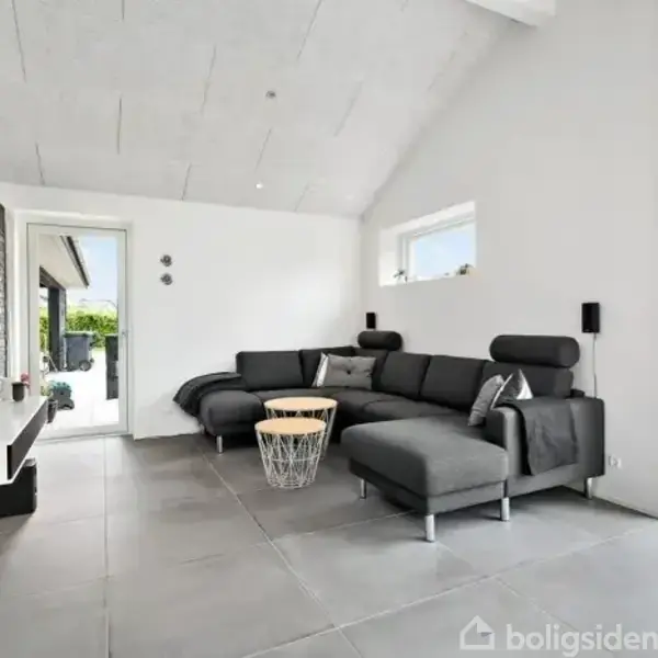 En moderne stue med en mørkegrå sofa placeret i hjørnet ved en væg. Der hænger et TV på den modsatte mur. Gulvet er dækket med grå fliser. Ved siden af sofaen står et rundt sofabord. Rummet er lyst, med store vinduer ud mod en have.