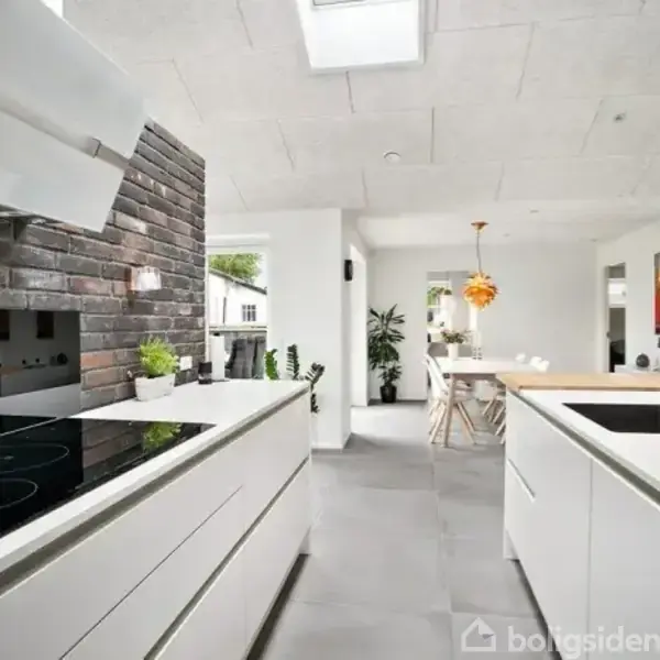 Moderne køkken omgivet af hvide skabe og grå fliser. En kogezone er ved en murstensvæg, og der er udsigt til en spiseplads med hængende lampe i baggrunden.