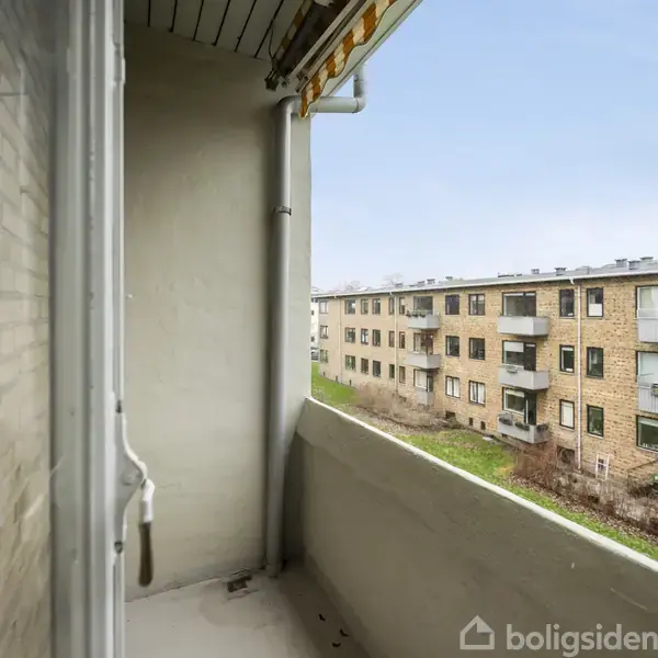 En lille altan med rækværk, der vender ud mod en gårdhave. Der er udsigt til flere beboelsesejendomme i mursten. Himlen er lys og overskyet.