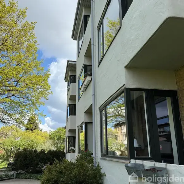 En moderne lejlighedsbygning med store vinduer står stille i en frodig have med grønne træer og buske omkring. Himlen er delvist dækket af hvide skyer.