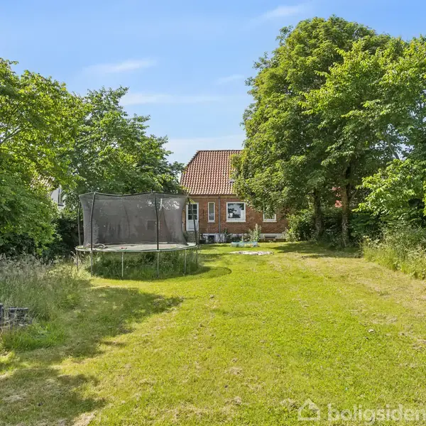 En have med en trampolin står på en græsplæne foran et rødt murstenshus. Omkring huset er der frodig grøn bevoksning og træer.