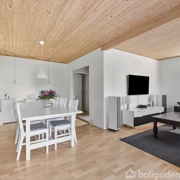 Spisestue og opholdsstue med hvide møbler og trægulv. Et spisebord med blomster i vase står nær vinduet. Sofa og sofabord med tv på væggen i baggrunden.