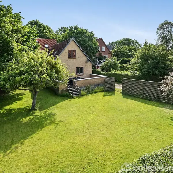 Et hus med tagvindue står foran en stor, velplejet græsplæne. Træer omkranser området, og en terrasse er synlig bag huset. Solrig dag.