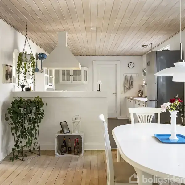En lys spisestue har et hvidt bord med blomster og en blå bakke, omgivet af hvide stole. Køkkenet i baggrunden har en bar, planter og et industrielt køleskab.