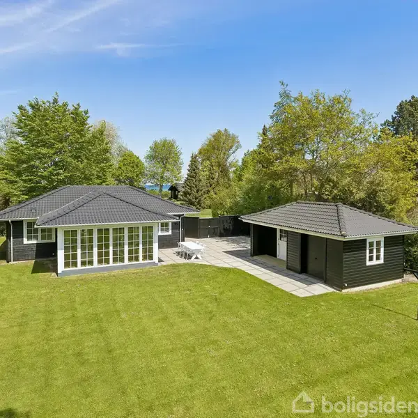 Et sortmalet sommerhus står stille omgivet af en grøn græsplæne og tæt skov. Til højre ses en mindre bygning. Skyfri blå himmel og sollys markerer en fredfyldt dag.