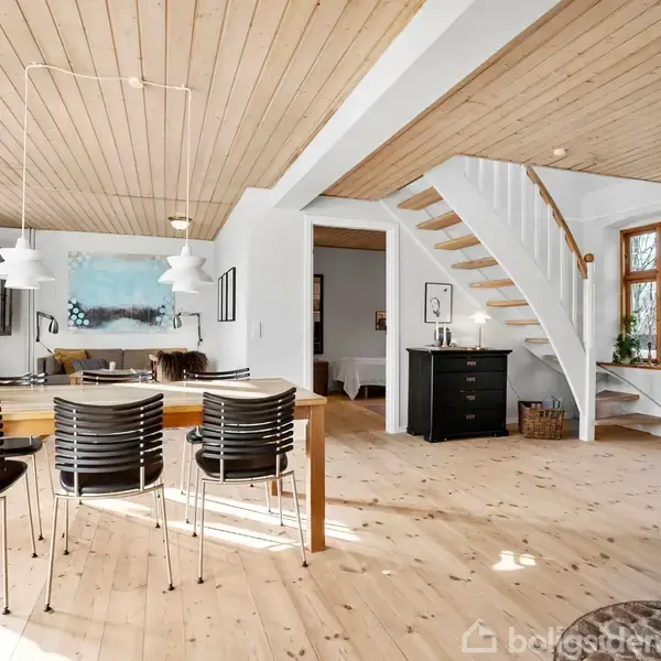 En spisestue med træbord og seks sorte stole; over bordet hænger tre hvide loftslamper. I baggrunden en stue med sofaer. Trætrappe fører til øverste etage. Lys træindretning.