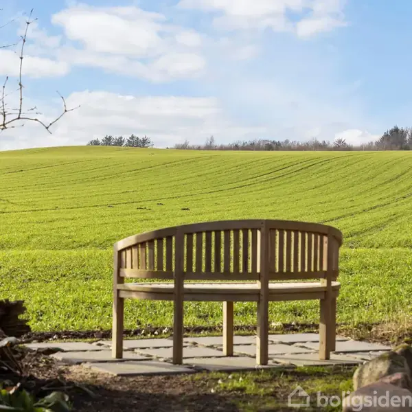 Bænk står stille på en lille flisebelagt plads, omkranset af træer, med udsigt til en grøn mark under en blå himmel med skyer.