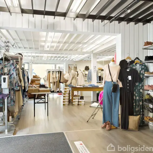 Mannequin udstiller tøj i en butik med hylder fyldt med sko og tøj. Rummets lyse interiør har et loft med lysarmaturer og flere tøjstativer i baggrunden.