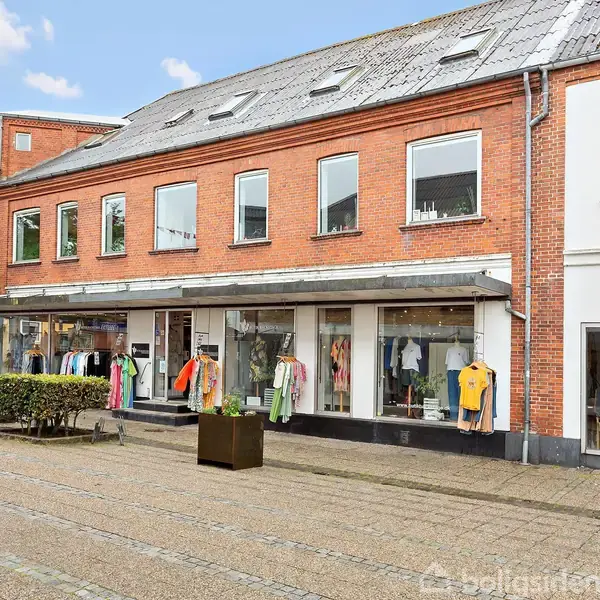 To-etagers butik med tøj på stativer udstillet udenfor, placeret på en gågade med fliser og træer. Sneklædte tags og brede vinduesfacader supplerer den røde murstensbygning.