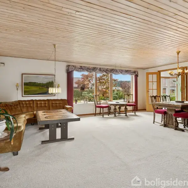 Stue med brun hjørnesofa og bord samt spisebord med røde stole, stående på hvidt tæppe. Store vinduer giver udsigt til en have. Loftet er af lyst træ.