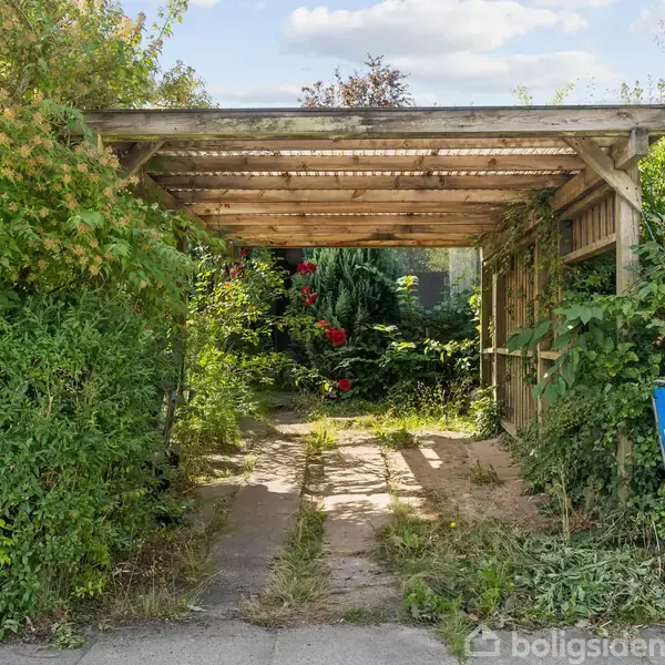 En carport, delvist dækket af planter, står ved en indkørsel. I baggrunden ses en frodig have. Et skilt på højre side siger: "Ny bolig? 262 817 83".