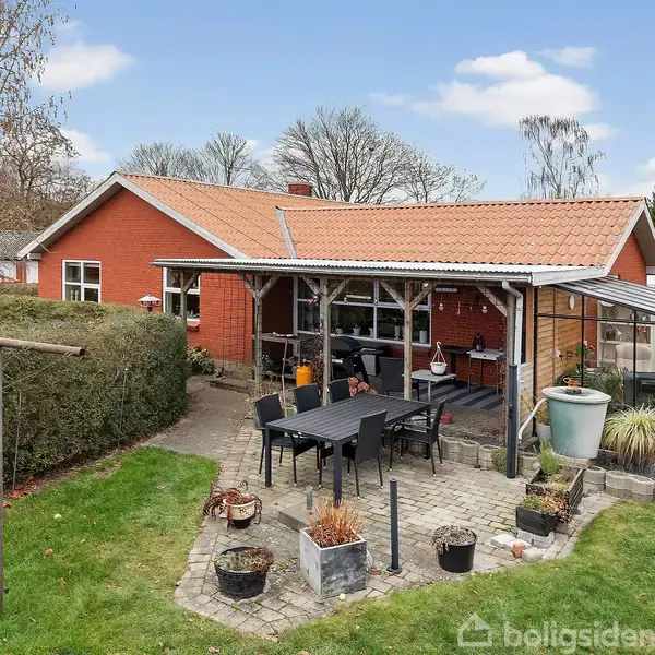Rødt murstenshus med terrasse i haven. En overdækket spiseplads og grønne planter omgiver terrassen. Baggrunden er en grøn have med buskede hegn og en græsplæne.