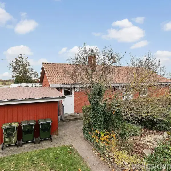 Rødt hus står stille omgivet af en have med en indkørsel ved siden af. Tre affaldscontainere er placeret ved garagen. Himlen er lysblå med spredte skyer.