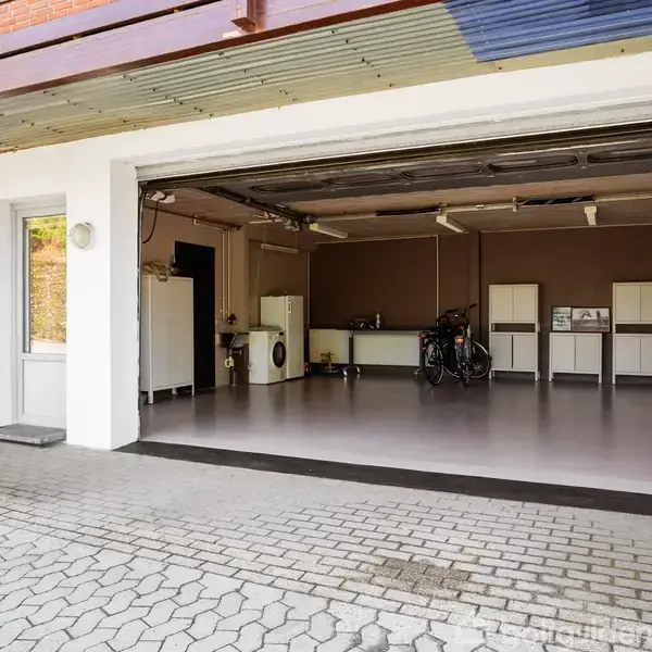 Garage med åbne døre, indeholder vaskemaskine, hylder og en cykel, placeret i en velholdt indkørsel med brostenslignende belægning og lyst interiør.