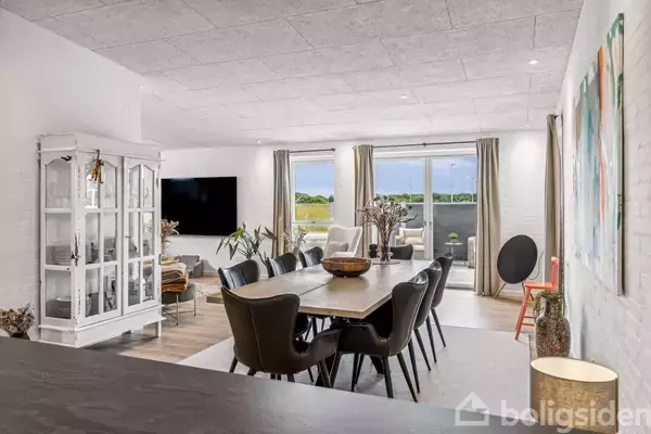 Spisebord med sorte stole står centralt i lyse stue. Store vinduer med gardiner åbner ud til en terrasse med grøn udsigt. Hvidt vitrineskab mod venstre. Ingen tekst.