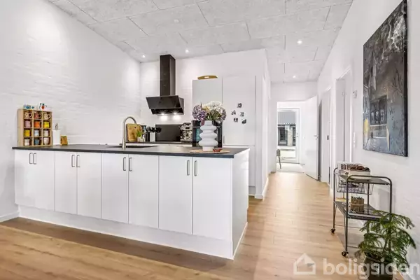 Et lyst køkken med hvide skabe og mørk bordplade. En vase med blomster står på bordet. En gang strækker sig væk fra køkkenet med kunstværk på væggen.