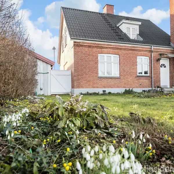 Murstenshus med sort tegltag og hvid rammedør står bag en frodig have med blomstrende erantis og vintergækker. Blå himmel og buske omgiver stedet.