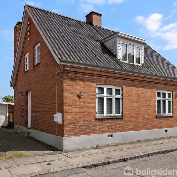 Rødt murstenshus med sort tag står stille i en rolig byzone. Grusvej og fortov omgiver huset; en garage og skraldespande ses til venstre. Klar blå himmel danner baggrund.