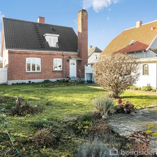 Et rødt murstenshus med sort tag står stille i en have med græsplæne og buske. En mindre bygning ses til højre, omgivet af fliser og planter.