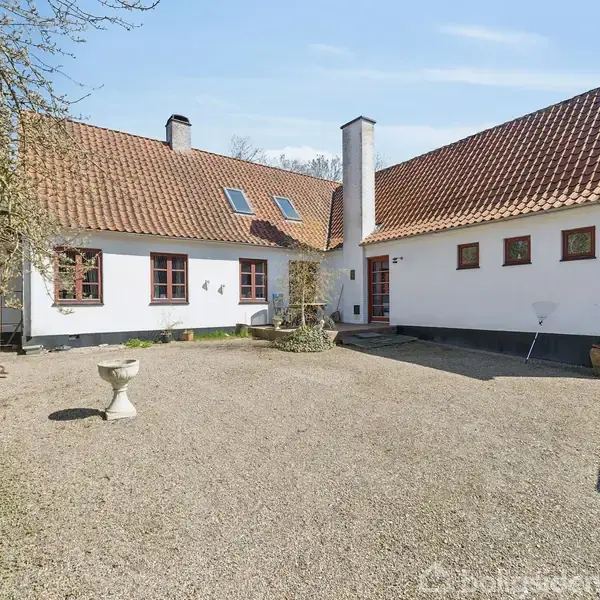 Hus med hvid facade og rødt tegltag står omkring en gårdsplads med grus. Der er flere vinduer, buskads omkring og blå himmel i baggrunden.