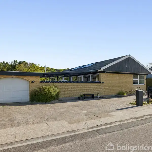 Hus med fladt tag står stille ved vejen, omgivet af have og indkørsel. Gul murstensfacade med hvid garageport til venstre. Omgivet af hække og have. Solrig dag.