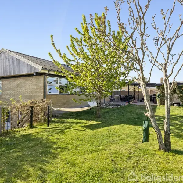 Et murstenshus står i en grøn have med græsplæne og nøgne træer. En hæk og et hegn omgiver området, mens en terrasse er synlig ved siden af huset.