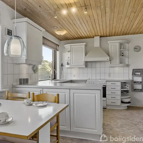 Et lyst køkken med hvide skabe og en emhætte over komfuret, omgivet af træloft og store vinduer med udsigt til naturen. Bordet har to kopper og en avis.