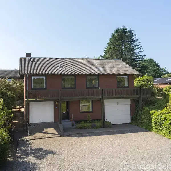 En to-etagers murstensvilla med to garager er omgivet af en velplejet have. Villaen står i solskin og er placeret i et grønt, beboet område med træer og anden beplantning.