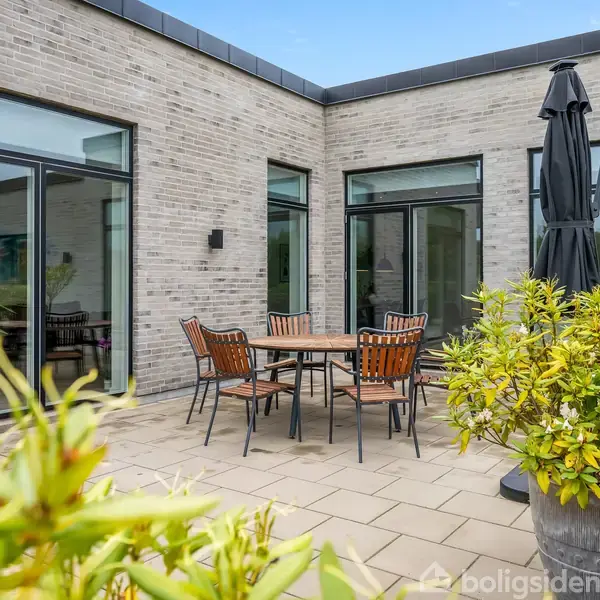 Rundt spisebord med stole står på flisebelagt terrasse. Parasol rejser sig ved siden af bordet. Omgivet af træer og planter. Murstensvægge med store vinduer danner baggrunden.