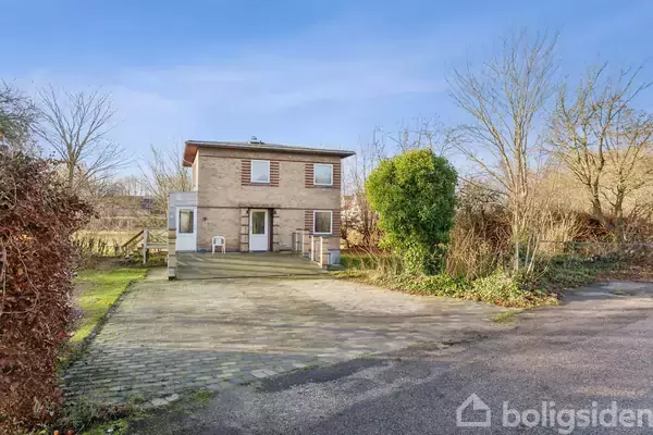 Et to-etagers hus står stille i en åben, haveomgivet kontekst med flisebelagt indkørsel og spredt bevoksning. En stol er placeret ved husets facade.