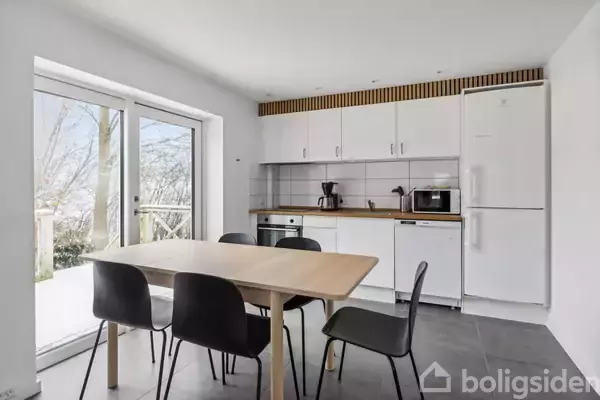 Et lyst køkken med et træbord omgivet af fire sorte stole. Der er hvide skabe, en ovn, kaffemaskine, mikroovn og et køleskab. Store vinduer viser udendørs natur.