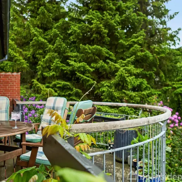 Rundt havebord med polstrede stole står på en terrasse. Baggrunden består af frodige træer og blomsterbuske, der indrammer den hyggelige udendørs scene.