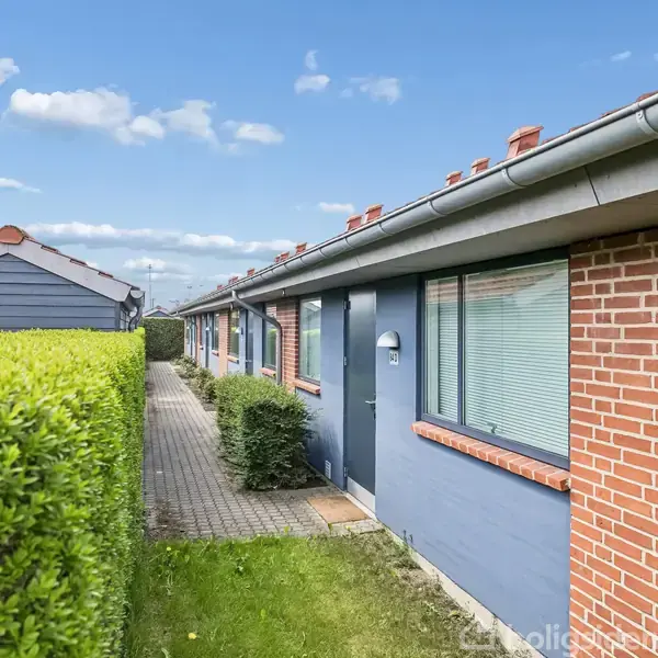 Rækkehus med røde mursten og blå facade ligger ved en stenbelagt sti omkranset af grønne hække, under en blå himmel med lette skyer.