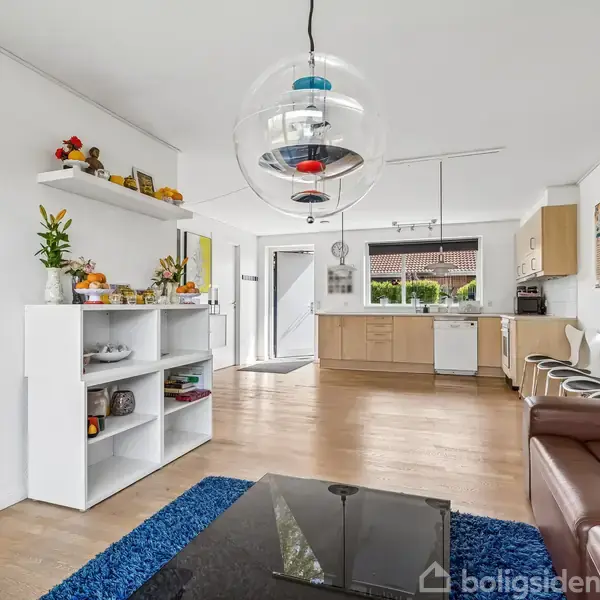 Rum med hvid bogreol fyldt med dekorationer, foran en brun sofa. I baggrunden ses et åbent køkken med træskabe og spiseområde. En stor, moderne lysekrone hænger fra loftet.