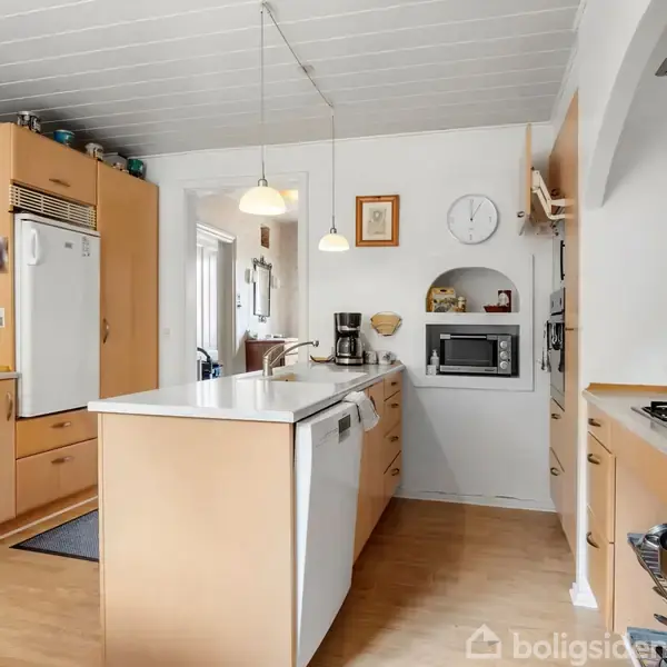 Køkkenet har træskabe og en hvid opvaskemaskine, med hængende lamper over bordpladen. I baggrunden er der et køleskab, mikroovn og en vægur. Toaster og vandkoger står på bordet.