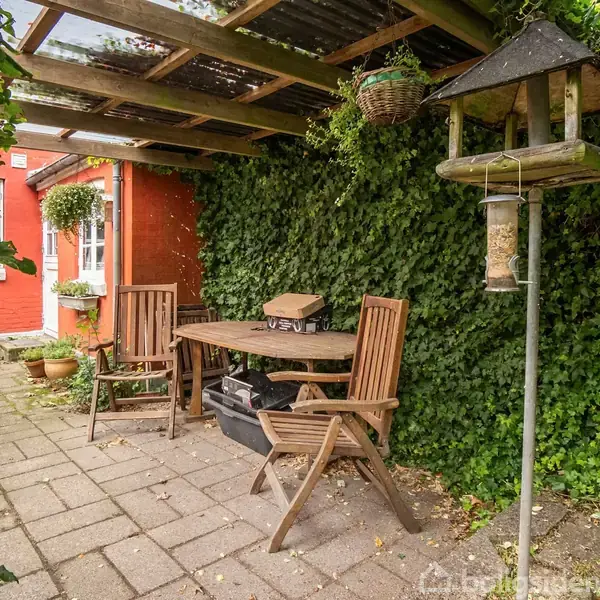 En træterrasse omfatter to stole og et bord under et halvtag, omringet af grønne planter. Ved siden af står et fuglehus på en stang. En rød murstensbygning ses i baggrunden.