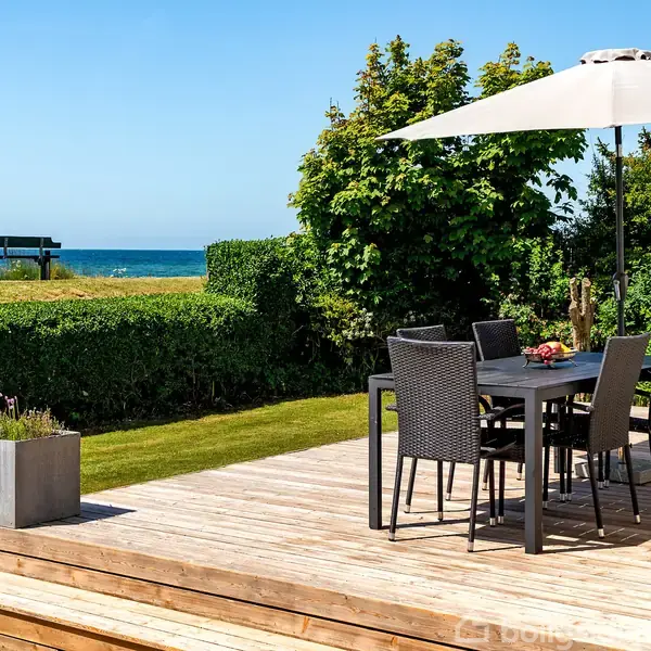 Terrasse med sort spisebord og stole står under parasol. Grøn græsplæne omgiver området, med buskads og træer. I baggrunden ses havet og en bænk. Ingen tekst fundet.