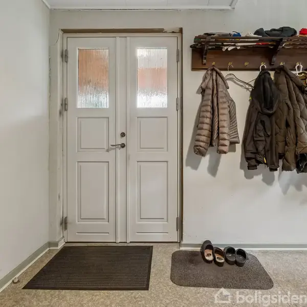 Dobbelt dør står lukket i en entré, flankeret af en knagerække med jakker til højre. To par sko står på måtter på gulvet under jakkerne.