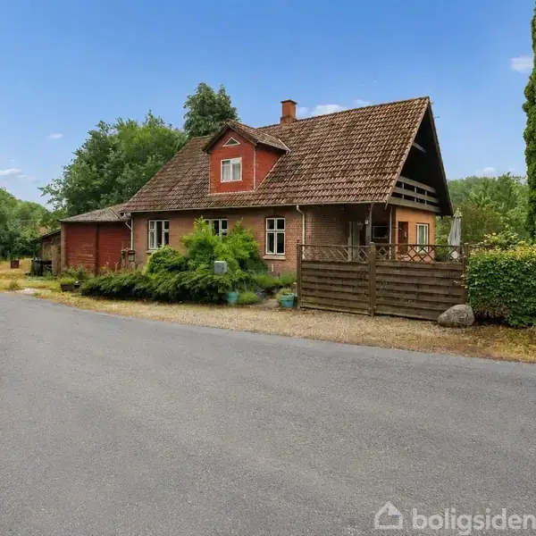 En rød murstenshus med skråtag står ved en vej i en frodig, grøn omgivelser. Huset har et åbent overdækket område med en træhegn foran.