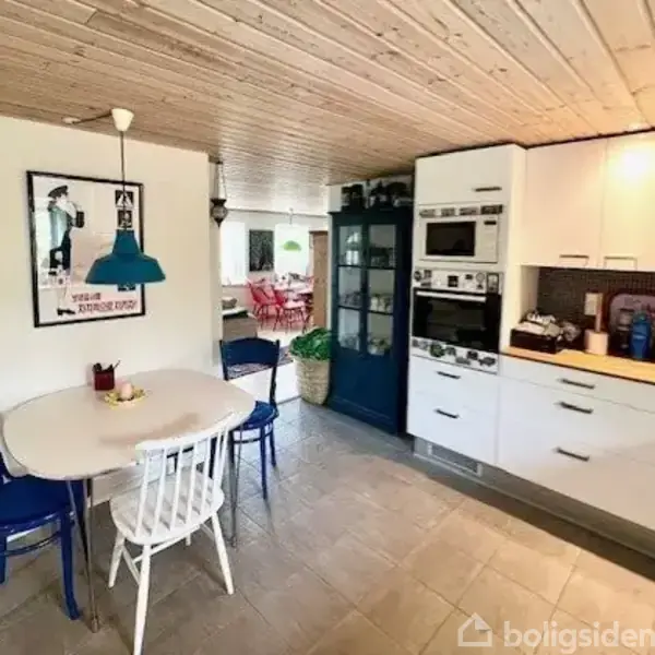 Et køkken med hvide skabe, indbyggede ovne og en lille spisekrog med et rundt bord og blå stole. Træloft og en mørk blå glasvitrine tilføjer til den hyggelige atmosfære.