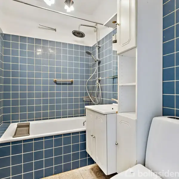 Et badeværelse med et hvidt badekar og bruser, omgivet af blå fliser. Der er en hvid vask med skab til højre og et toilet ved siden af.