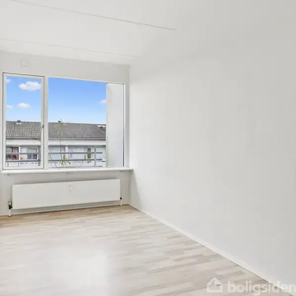 Et tomt rum med lyse trægulve, hvor en radiator står under et stort vindue. Udsigten viser nabobygningens tag og blå himmel. Hvide vægge omgiver det enkle rum.