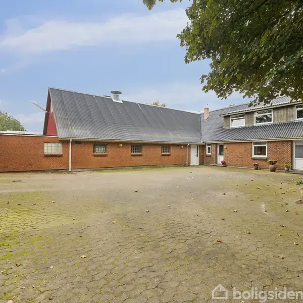 Et gårdområde med brostensbelægning omgivet af røde murstensbygninger med skrå tage. En stor træ står til højre, og himlen er lys og klar.