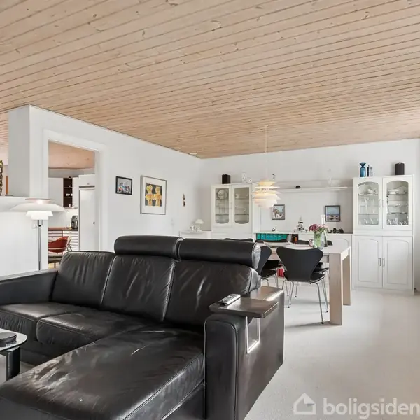 Sort lædersofa står i et lyst stueområde med træloft. Sofaen vender mod et lille sofabord. I baggrunden ses spisebord, reoler og billeder på væggene.