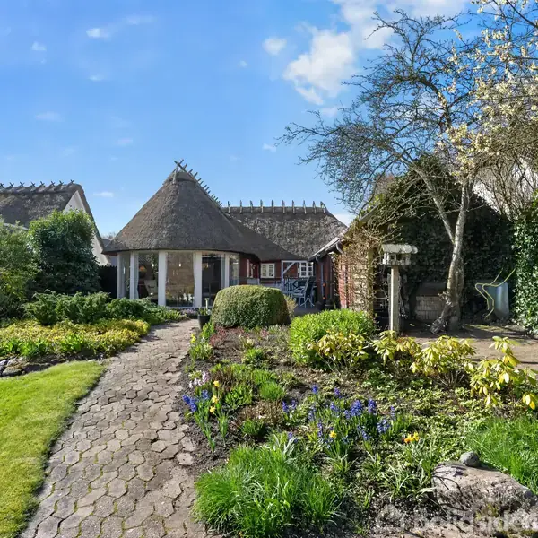 Rundt stråtækt hus med stor have fyldt med blomster og grønne planter. En stenbelagt sti leder ind til huset, omgivet af træer og buske, under en klar blå himmel.